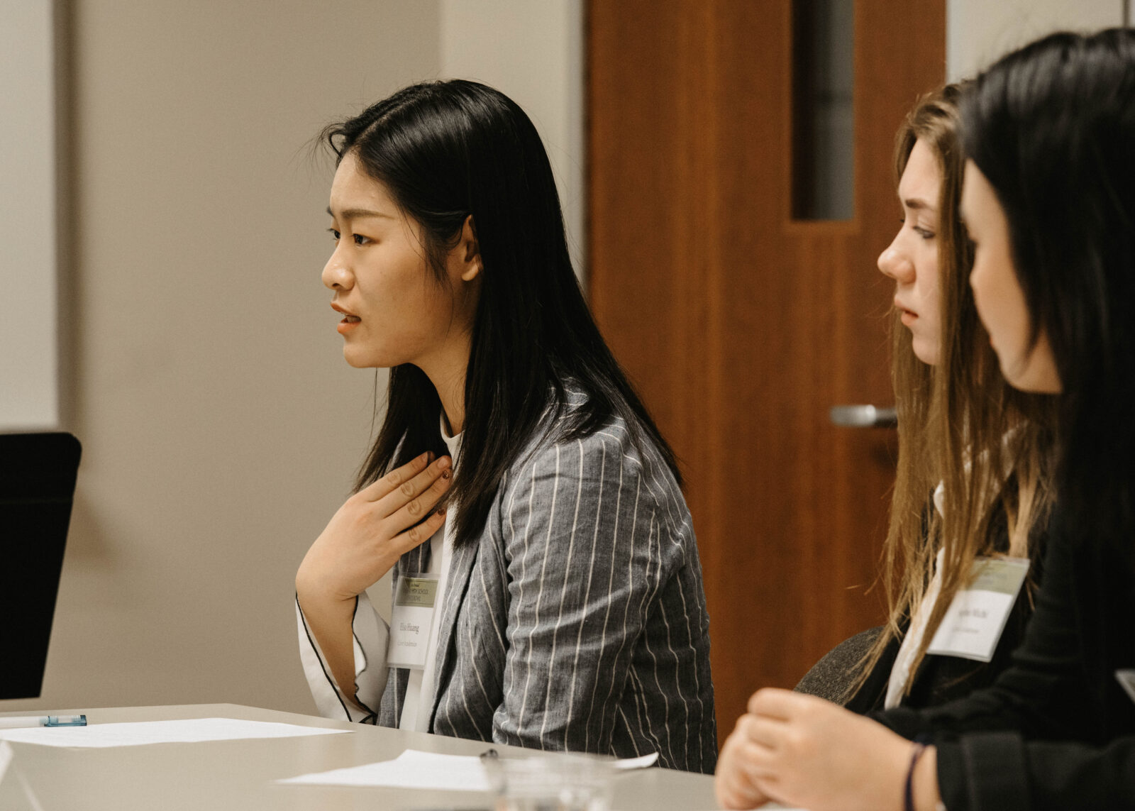 Photograph of a young woman speaking to a group out of frame. She is pointing to her chest while she makes a point. Two other young women are seated next to her and watch her speak.